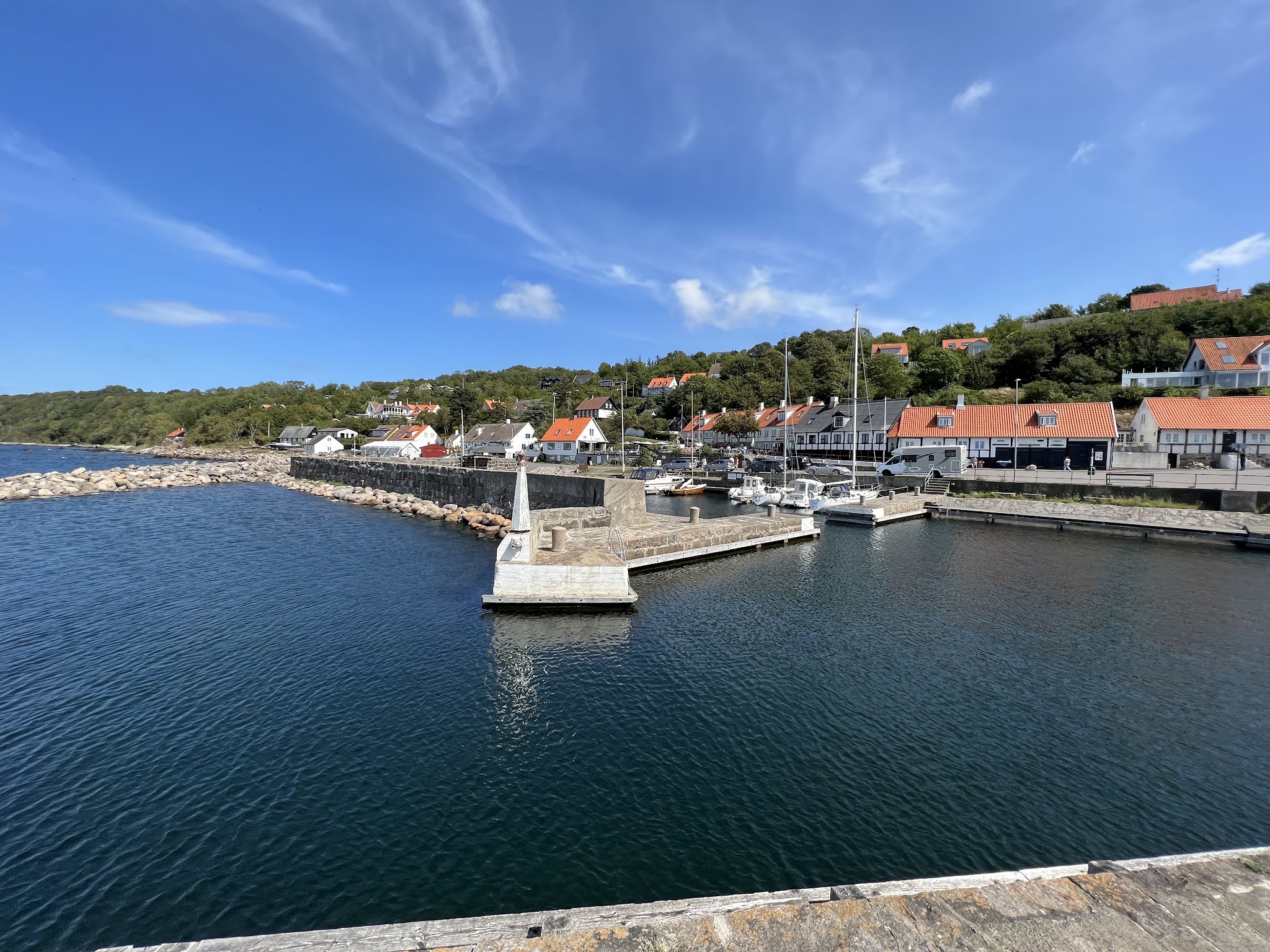 Kleiner Küstenhafen mit Segelbooten, Mole und roten Ziegeldächern vor grünem Hügel unter klarem, blauem Himmel.