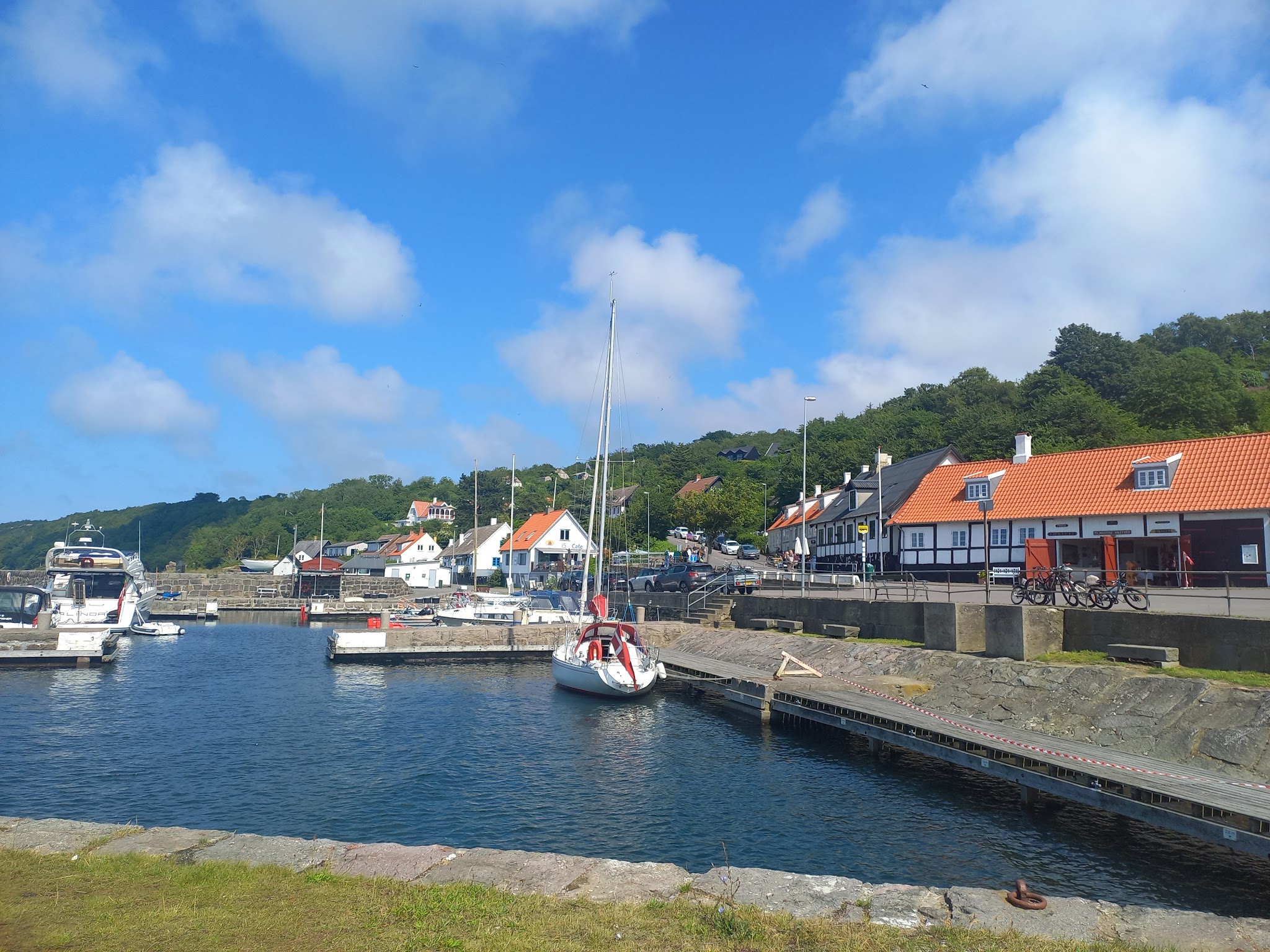 Kleiner Küstenhafen mit Segelbooten, weißen Häusern mit orangefarbenen Dächern, umgeben von grüner Hügellandschaft