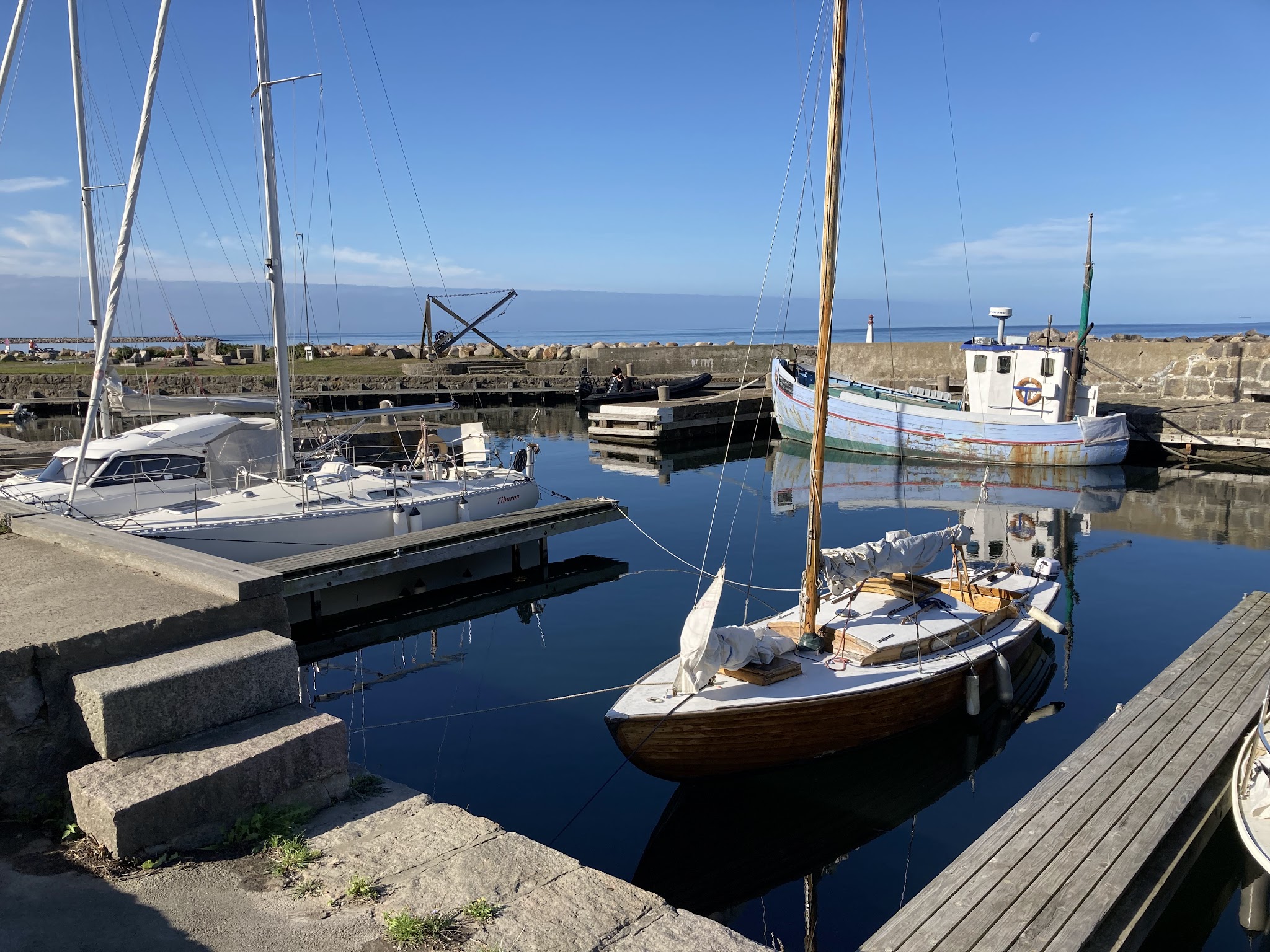 Kleiner Hafen; Holzsegelboot vorne, moderne Yacht und verwittertes Fischkutter spiegeln sich im ruhigen Wasser.