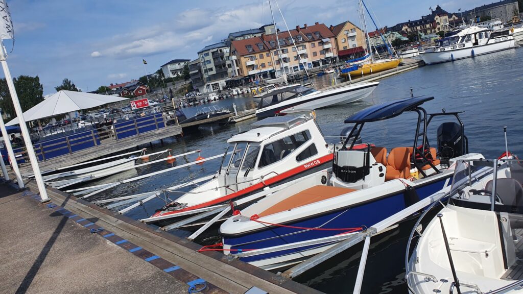 Mehrere Motorboote liegen am sonnigen Yachthafen; dahinter Uferpromenade mit Wohnhäusern und Cafés.