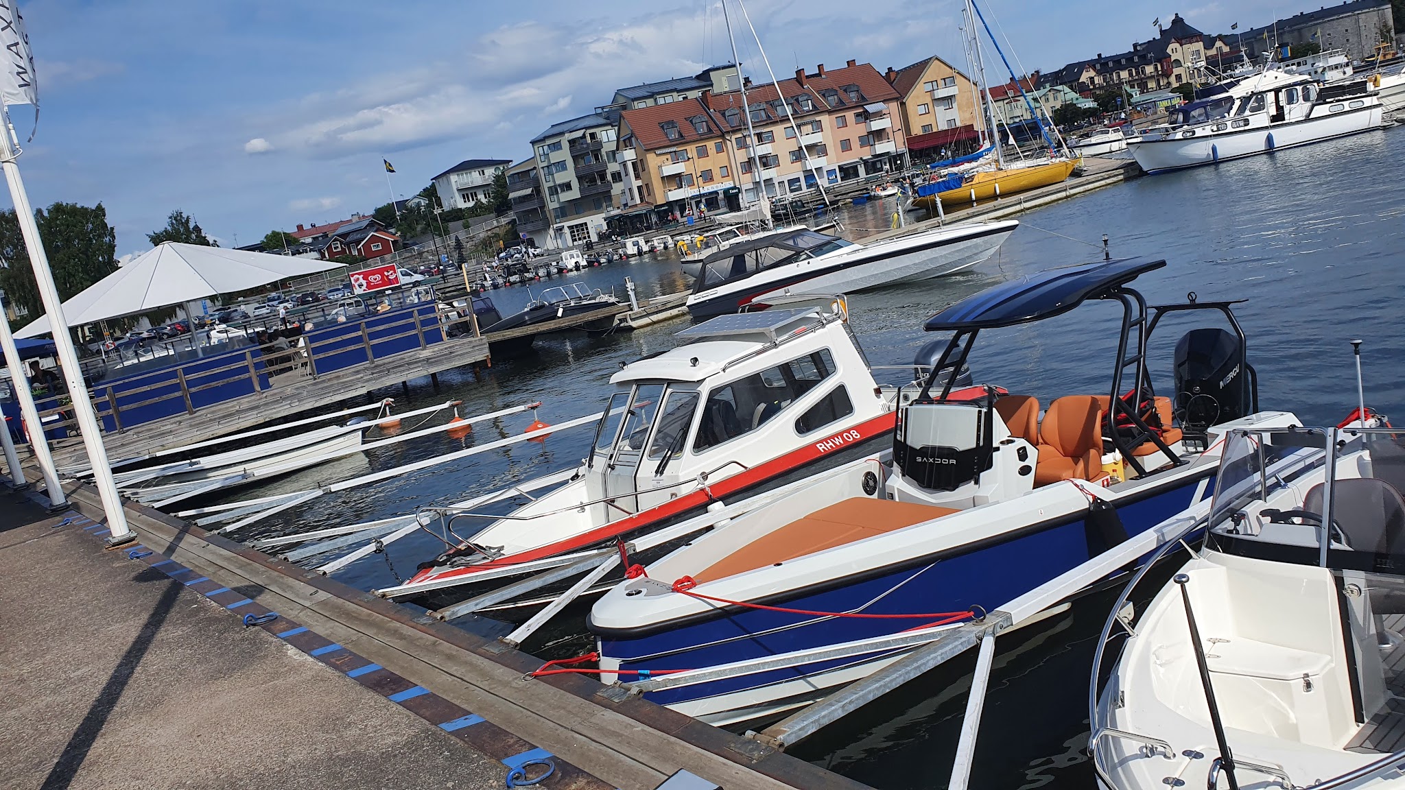 Mehrere Motorboote liegen am sonnigen Yachthafen; dahinter Uferpromenade mit Wohnhäusern und Cafés.
