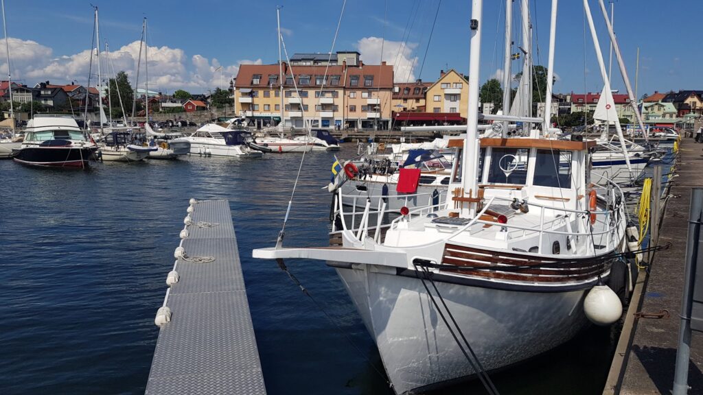 Weiße Boote im sonnigen Hafen, vorn ein Kajütboot mit schwedischer Flagge, Pier und Häuser im Hintergrund