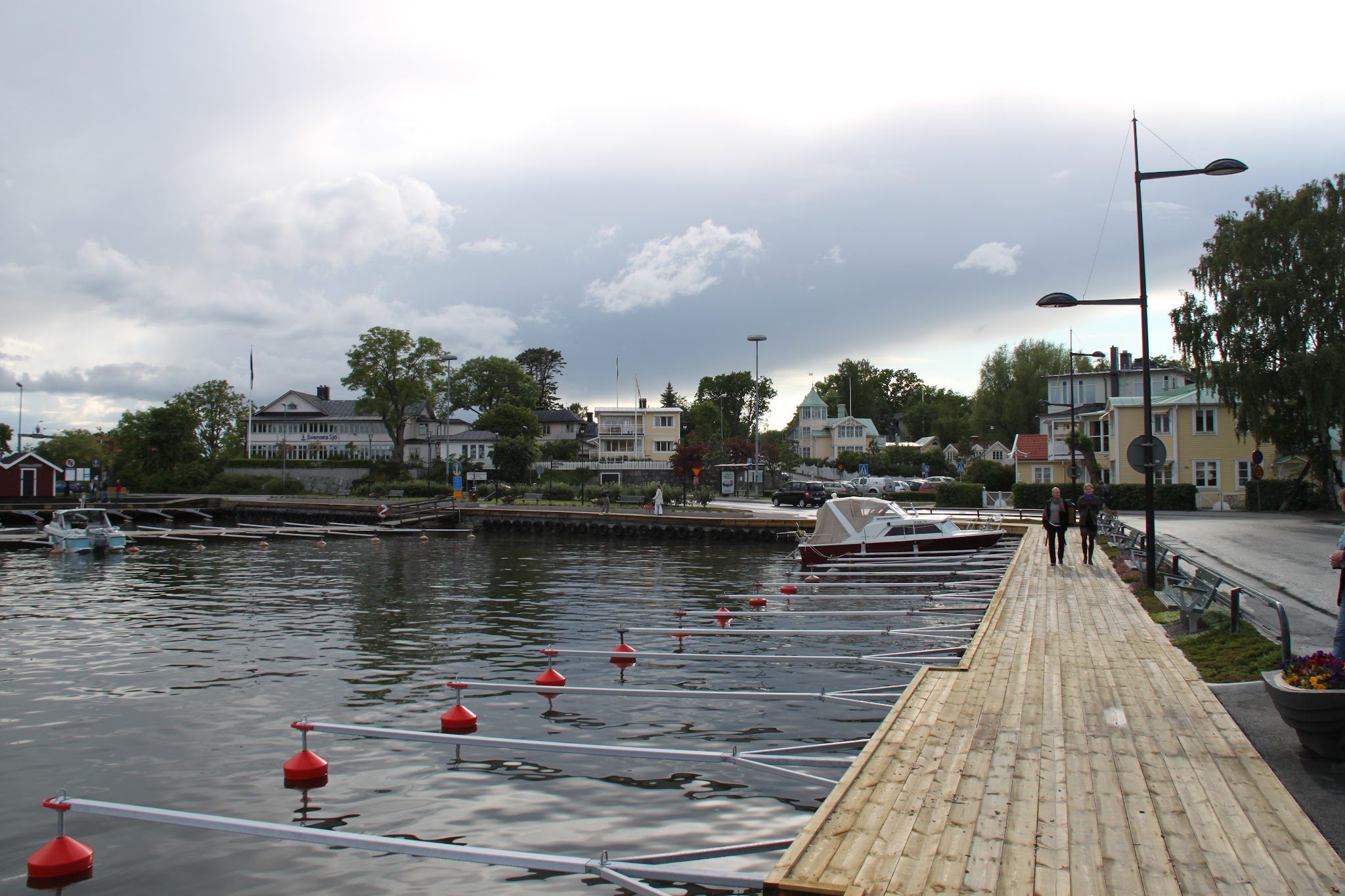 Holzsteg mit leeren Bootsplätzen und roter Boje, Motorboot, zwei Spaziergänger; dahinter Uferhäuser unter wolkigem Himmel.