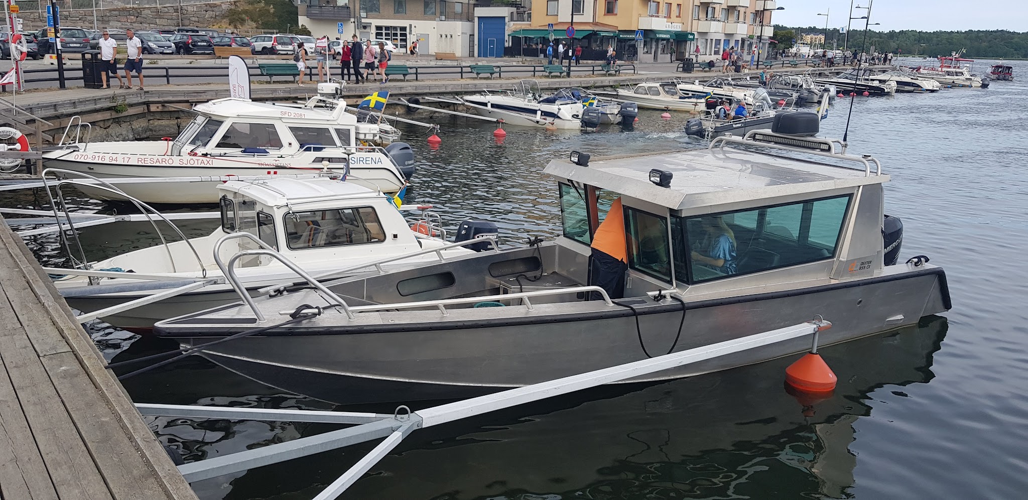 Mehrere Motorboote liegen im Stadthafen an, Menschen spazieren auf der Kaimauer, Häuser im Hintergrund.