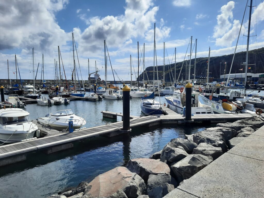 Marina mit vielen Segel- und Motorbooten an Stegen; felsige Uferkante vorn, bewölkter Himmel mit Sonne