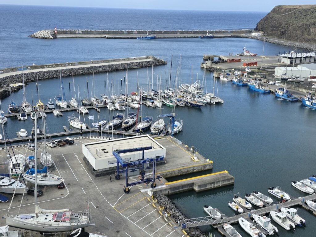 Kleiner Yachthafen mit vielen Segelbooten, Pier und Mole vor offenem Meer; felsige Steilküste rechts