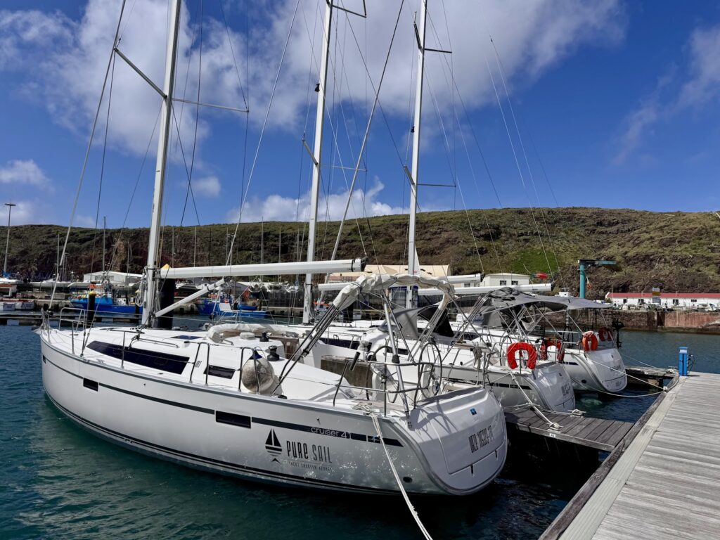 Mehrere weiße Segelyachten liegen an einem Steg im Hafen vor grüner Felsküste und blauem Himmel.