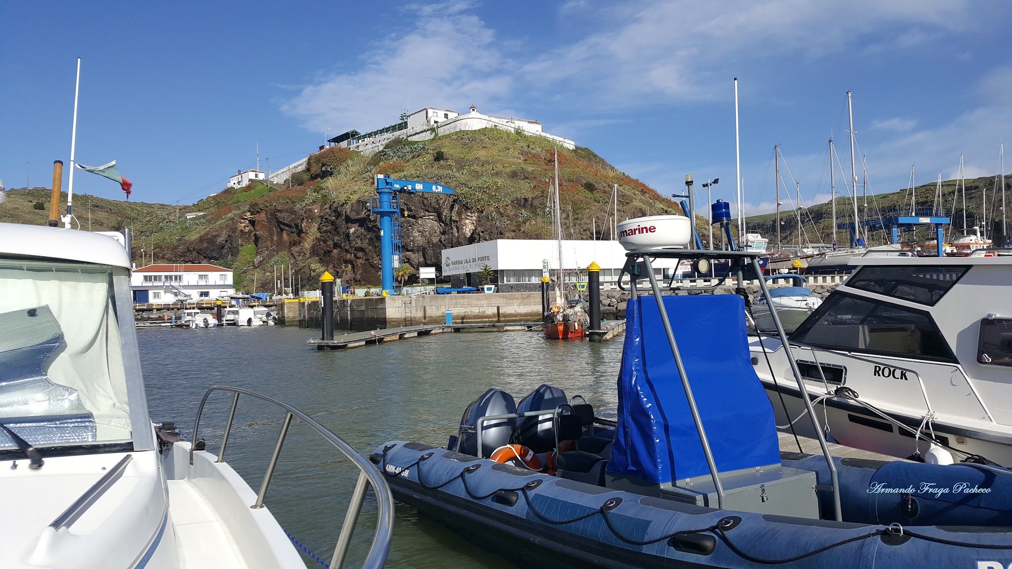 Boote im Hafen vor grünem Hügel mit weißer Festung unter blauem Himmel