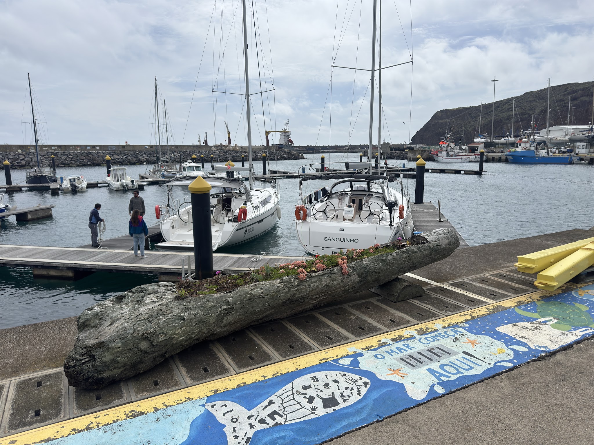 Segelboote im Hafen, drei Menschen auf Steg; bemalter Kai mit Meeresmotiv, Blumen in ausgehöhltem Baumstamm vorn