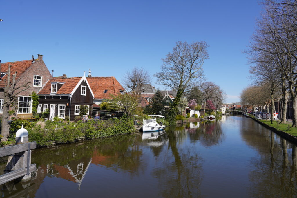 Ruhiger Kanal mit Boot, roten Ziegeldach-Häusern und blühenden Bäumen unter blauem Himmel