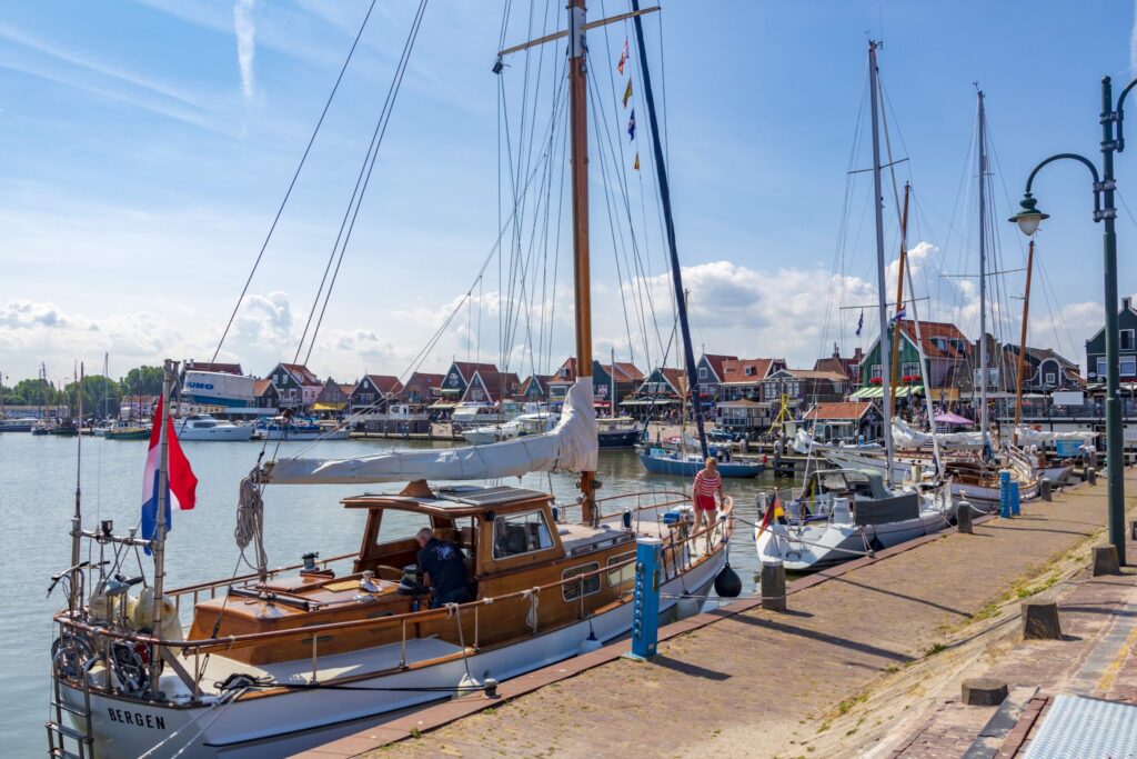 Hafen mit Holzsegelbooten am Kai, niederländische Flaggen, bunte Giebelhäuser und blauer Sommerhimmel.