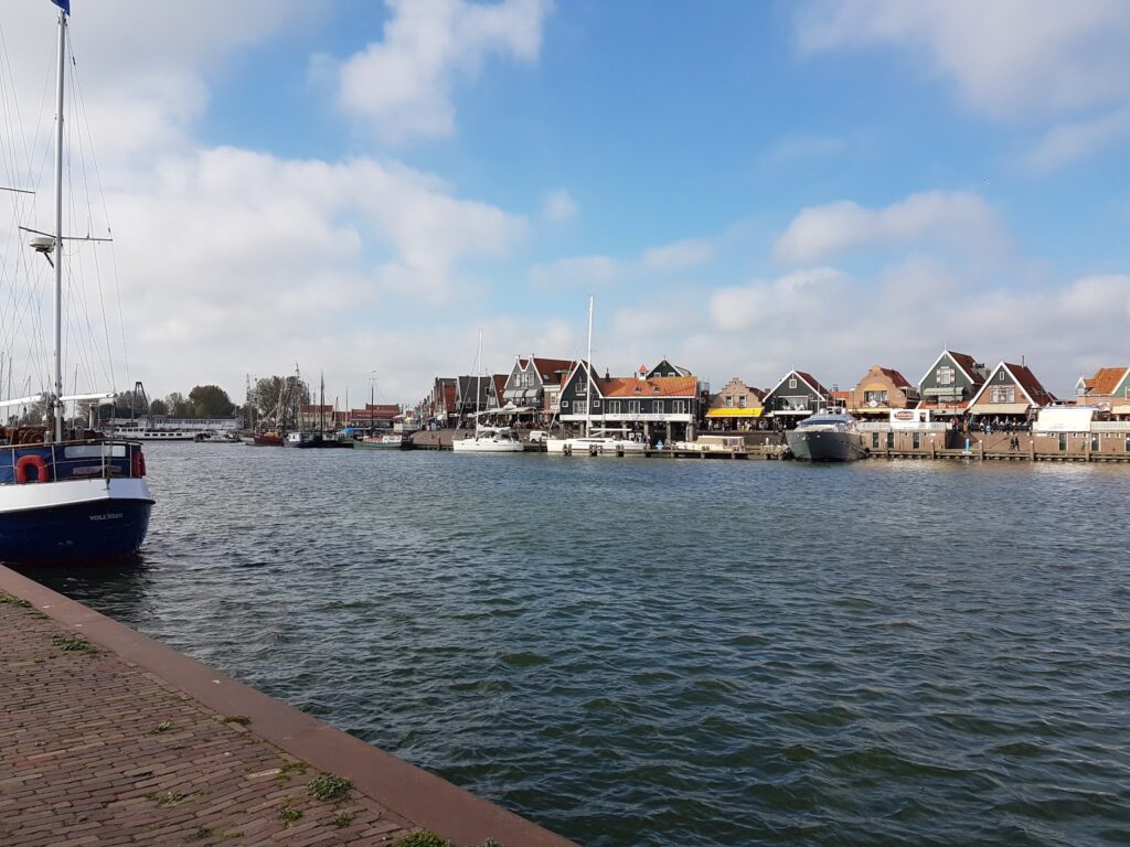 Kleiner Hafen: Segelboot links im Wasser, bunte Giebelhäuser und weitere Boote gegenüber unter blauem Himmel.