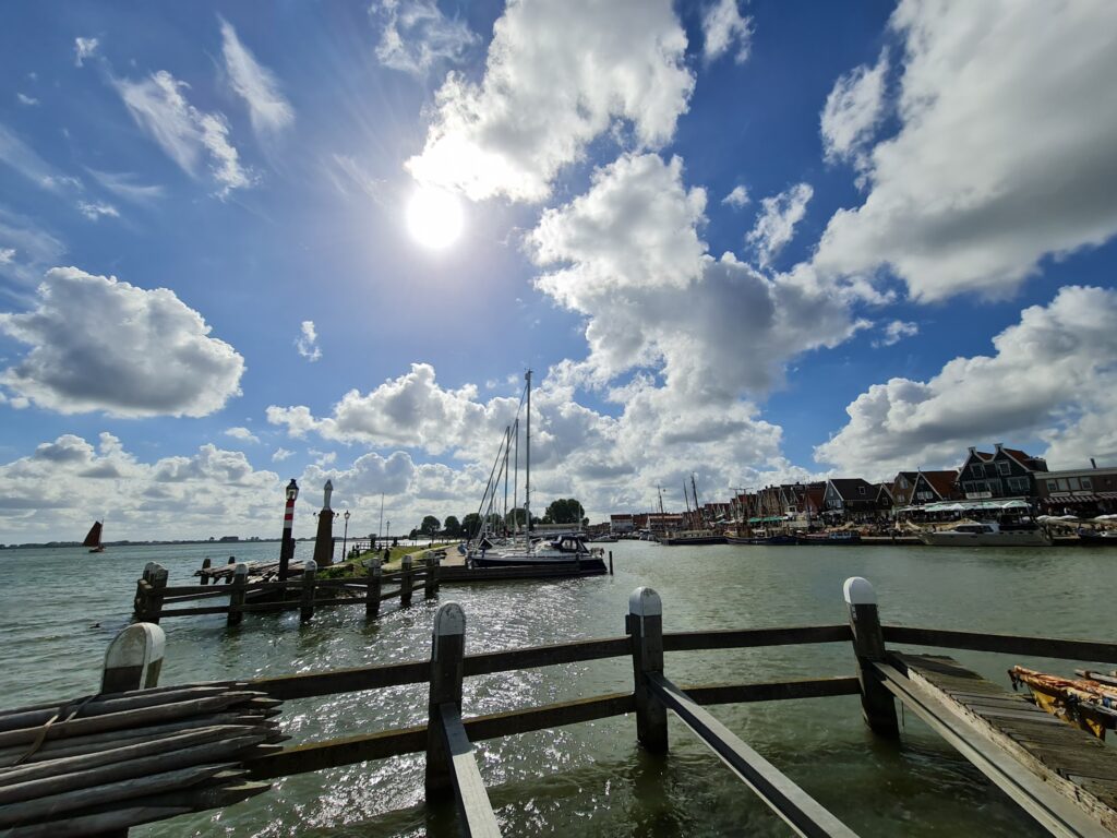 Sonniger Hafen: Holzböden vorn, Segelboote am Steg, dahinter Häuserzeile am Wasser unter blauem Himmel mit Wolken.