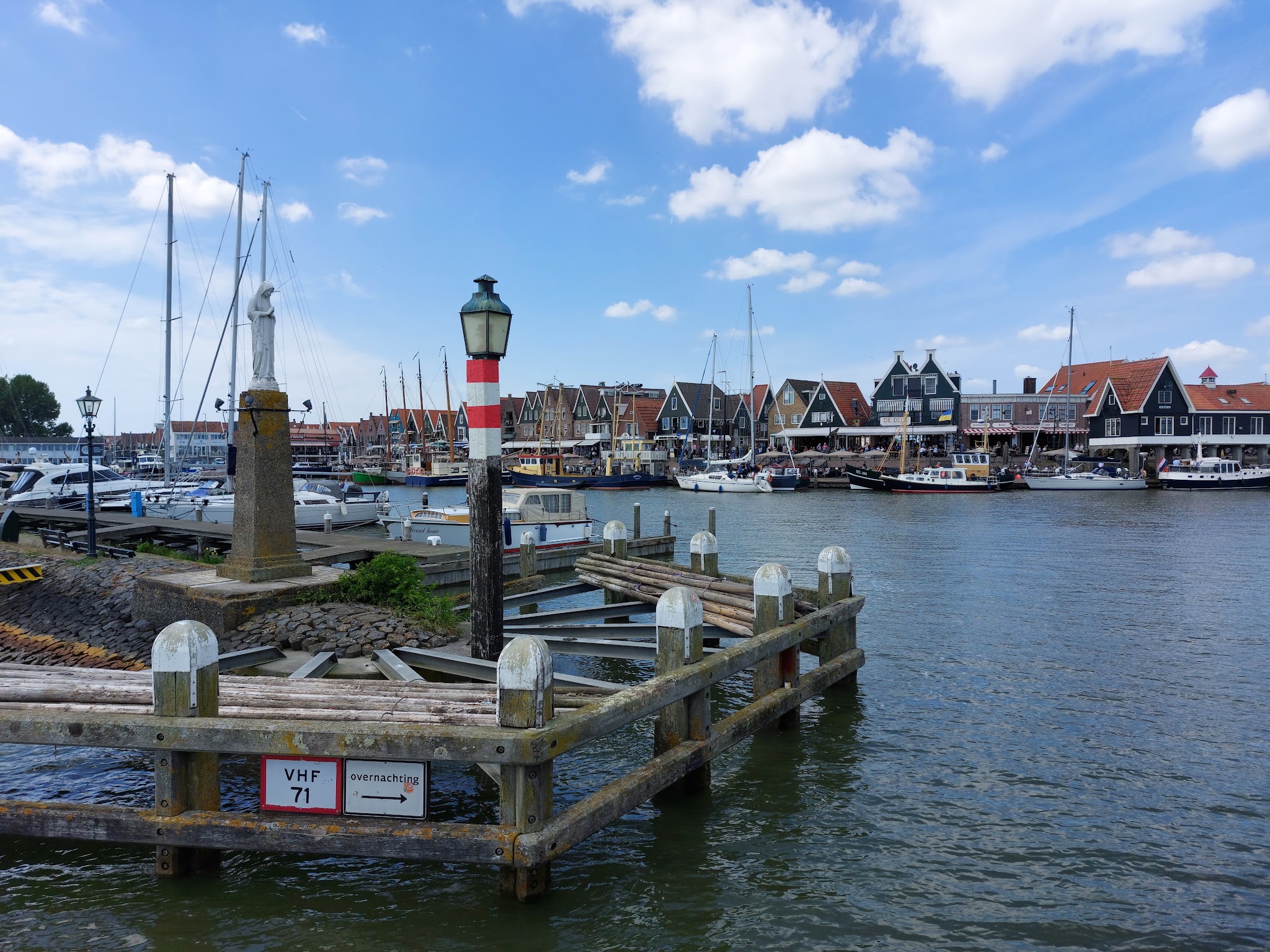 Hafen mit Segelbooten, historische Giebelhäuser, Holzsteg, Statue und rot-weißer Mast unter blauem Himmel