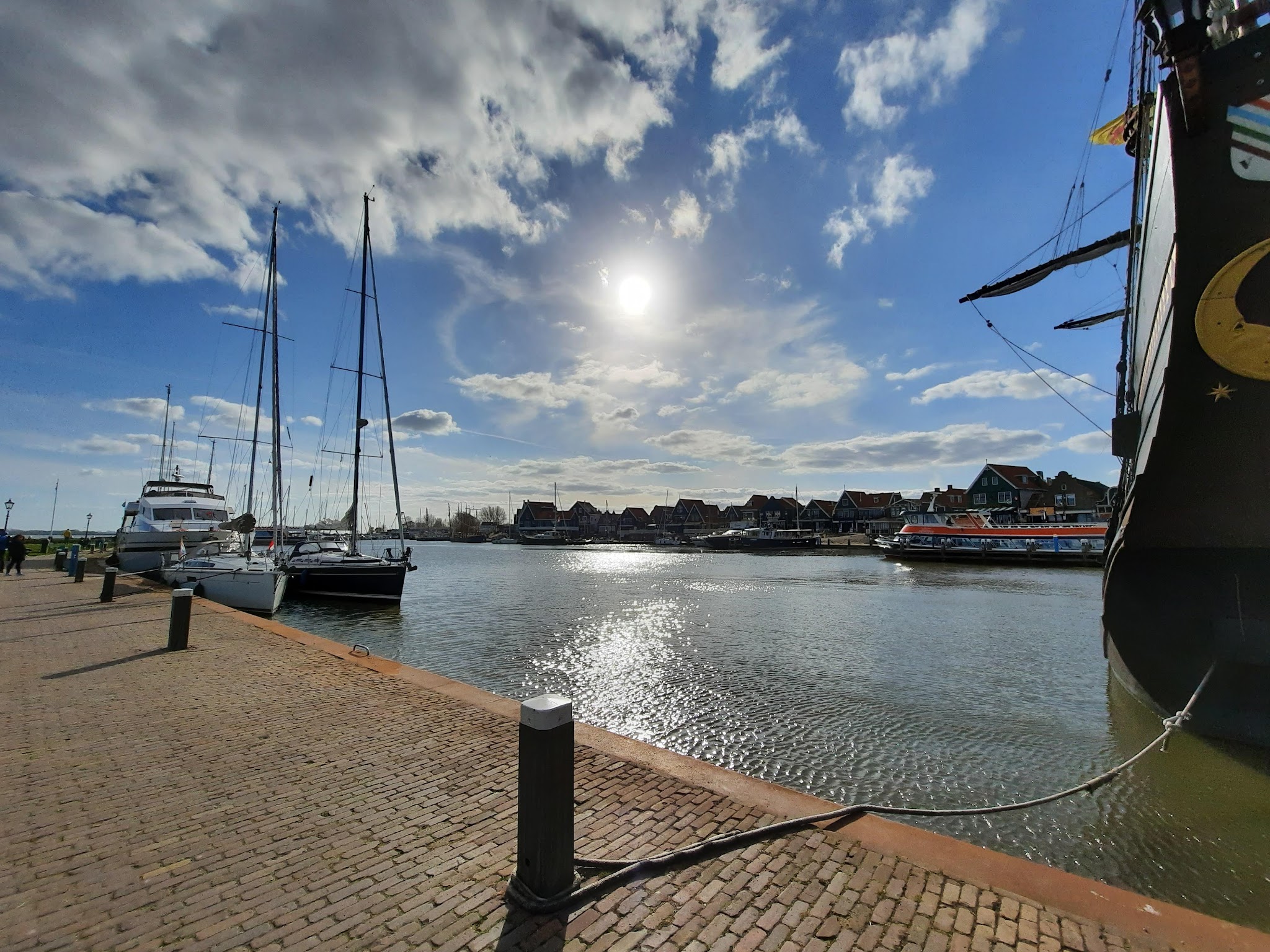 Sonniger Hafen: Segelboote am Steg, historisches Schiff rechts, Kopfsteinpflasterkai und Reihenhäuser im Hintergrund.