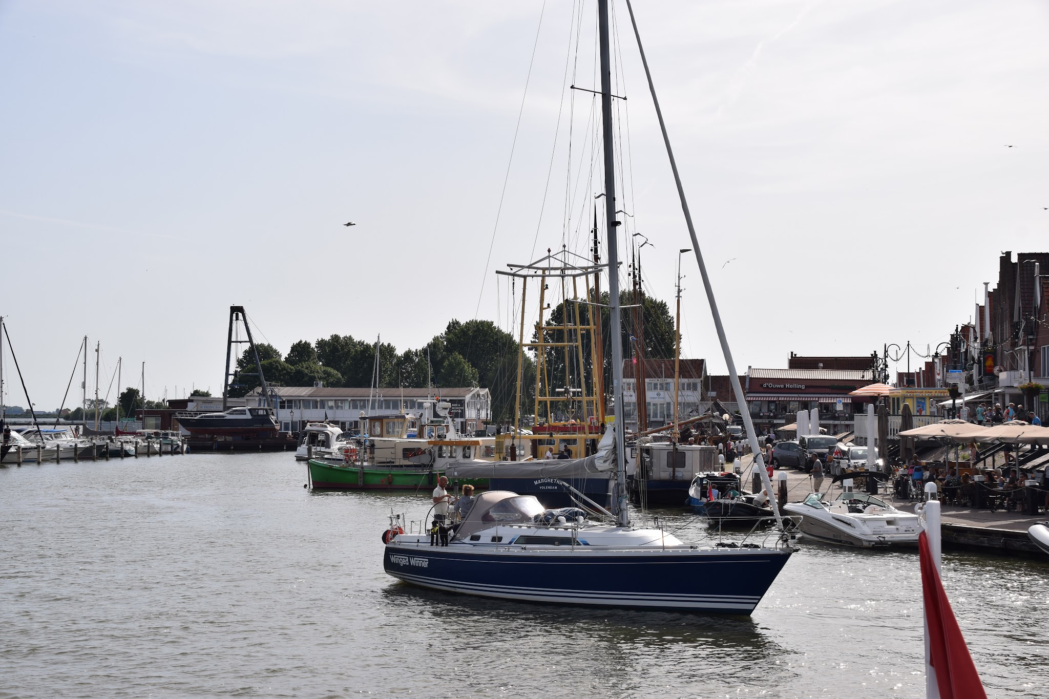 Segelboot fährt in belebten Yachthafen; Boote an Stegen, Menschen flanieren an Uferpromenade im Sommer.